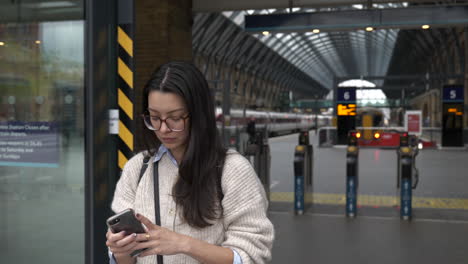 Una-Joven-Atractiva-Espera-Afuera-De-Una-Estación-De-Tren-Usando-Su-Teléfono-Inteligente-Móvil