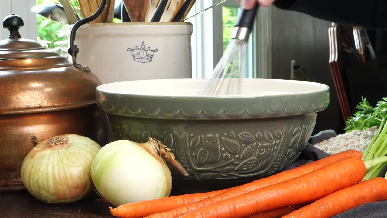 Making meatballs for Italian Wedding Soup. Using a whisk to mix ingredients in a big green ceramic bowl on a counter in the kitchen. Fresh vegetables like carrots and onions on the counter.