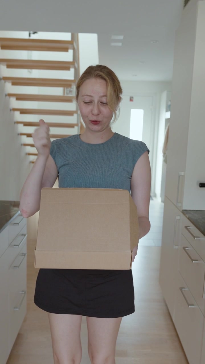 Donna sorridente che cammina con un pacco di cartone a casa, rappresentando la consegna, lo shopping online, la ricezione di pacchi e la soddisfazione del cliente.