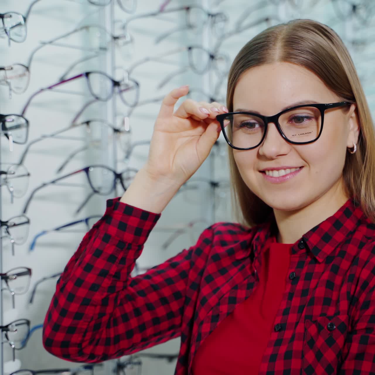 Beautiful girl in red shirt choosing new eyeglasses. Smiling attractive young woman holding stylish glasses in the optical shop.