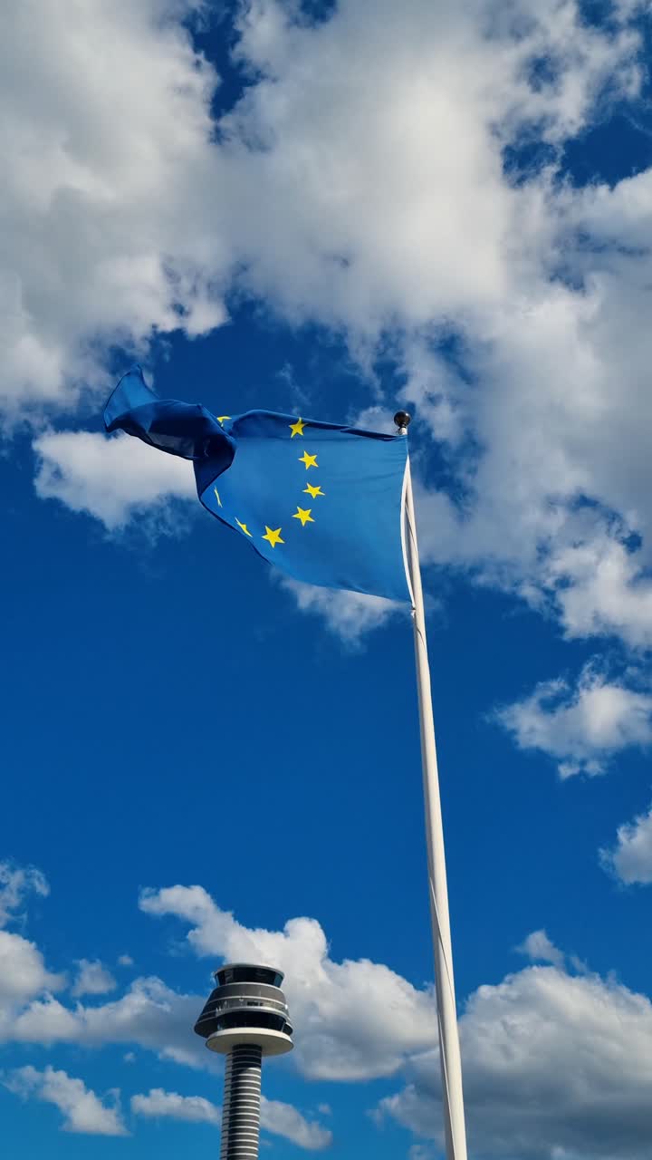 The European Union flag sways in the cold spring wind under a bright blue sky, its twelve golden stars symbolizing unity and harmony as white clouds drift slowly overhead, filmed in Arlanda, Sweden