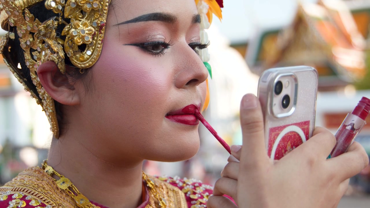 Close-up of a Thai classical dancer applying red lipstick while using a smartphone mirror