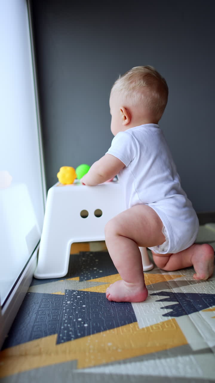 Small baby boy in white bodysuit stands near the chair. Lovely infant kid playing at home. Vertical video.