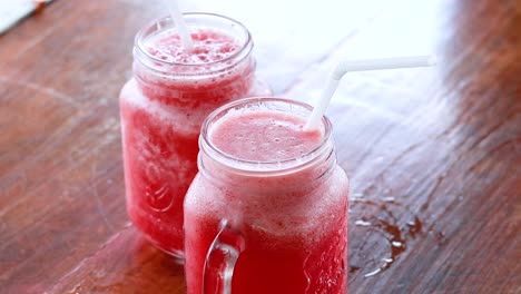 two glasses of watermelon juice on a wooden table, with hands raising one glass for a toast. bright natural lighting