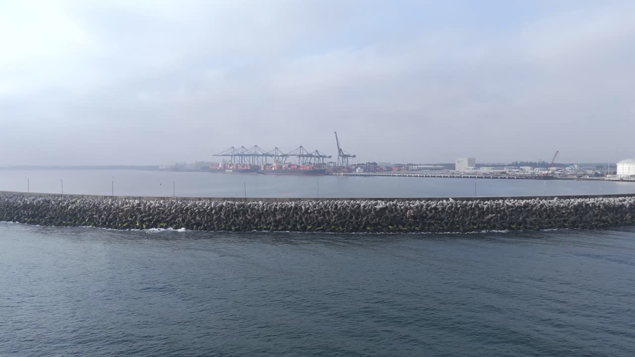 A panoramic shot of a busy container port, featuring massive cargo cranes and industrial buildings in the distance, under cloudy cold weather.