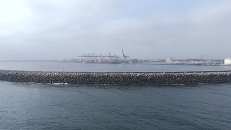 a panoramic shot of a busy container port, featuring massive cargo cranes and industrial buildings in the distance, under cloudy cold weather.