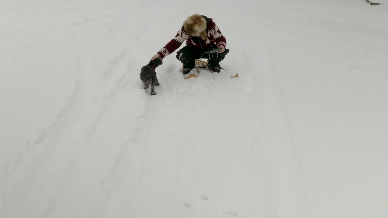 큰 모피 모자를 쓴 성인 남자, 캐나다 북부에 있는 애완견과 함께 얼어붙은 호수에 있는 전통적인 눈신을 신고 카메라를 향해 손을 흔들고 있습니다.