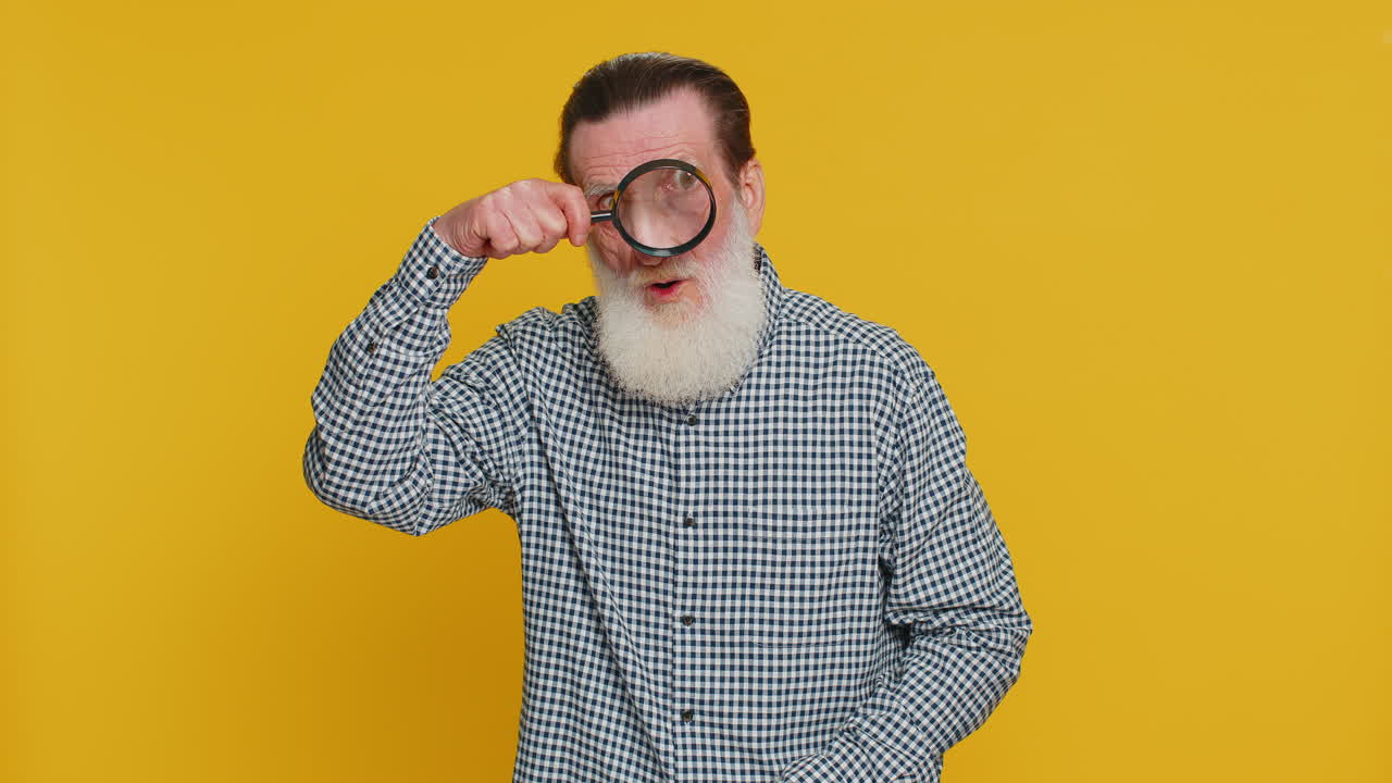 Senior old man holds magnifying glass near face looking at camera with big zoomed eye analyzing