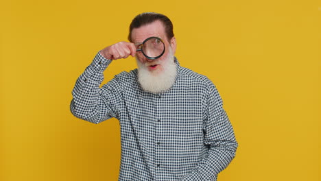 senior old man holds magnifying glass near face looking at camera with big zoomed eye analyzing