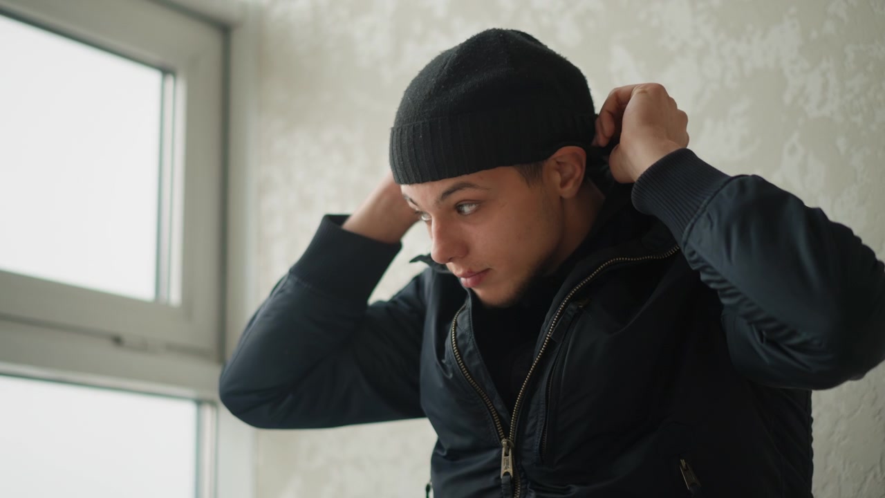 Close up of man wearing dark jacket and beanie adjusting clothing indoors near window. Man handles fabric with calm expression in softly lit room featuring light colored textured wall