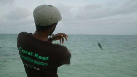 medium shot of a young black man throwing a stone into the sea