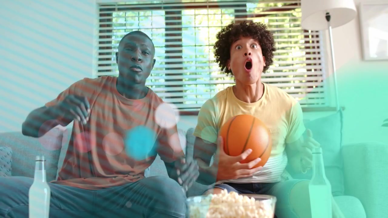Two men watching sports, TV play starting, holding basketball, eating popcorn, graphics covering
