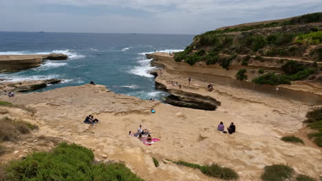 Toma-En-ángulo-Alto-Sobre-Las-Olas-Del-Mar-Salpicando-A-Lo-Largo-Del-Arco-De-Piedra-Caliza-En-Malta-Con-Turistas-Sentados-Disfrutando-De-La-Vista