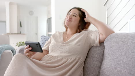 Glückliche,-Gemischtrassige-Plus-Size-Frau-Mit-Smartphone-Videoanruf-Im-Sonnigen-Wohnzimmer,-Zeitlupe