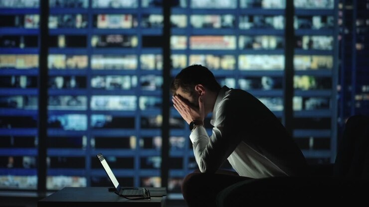 Tired young man working on a laptop late night in the office. Sleepy Businessman sitting at desk in dark office. Tired and stressed businessman in glasses works on a laptop of the night city office