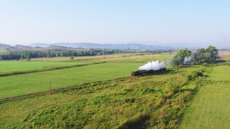 A cinematic, high aerial view from a drone follows a steam train on its journey. The historic locomotive travels alone through a vast, green rural landscape of fields and rolling hills