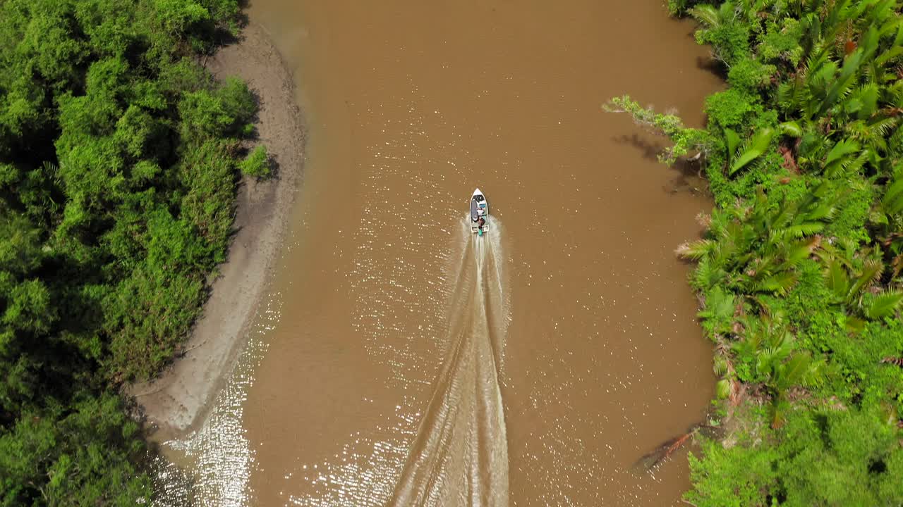 Aerial top down tracking shot of boat in murky river through lush green Indonesian jungle