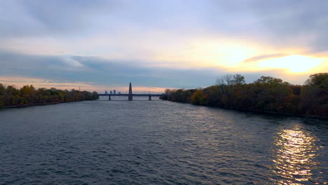 Hermosa-Vista-Dorada-De-La-Puesta-De-Sol-Del-Río,-Agua-Que-Fluye-En-El-Parque-Jean-Drapeau-Montreal,-Relajante-Paisaje-Natural,-Vegetación-árboles-En-Ambos-Lados,-Cielo-Nublado-Y-Puente-En-Segundo-Plano