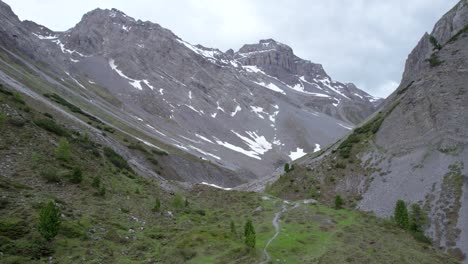 Aerial-drone-footage-rising-straight-up-to-reveal-a-glacial-mountain-landscape-with-patches-of-snow,-isolated-trees-a-remote-alpine-hiking-trail-in-Switzerland