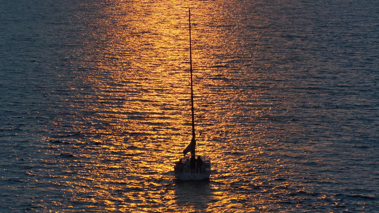Aerial drone view of a sailboat gliding on golden, sunlit water during sunset, with shimmering reflections surrounding the vessel