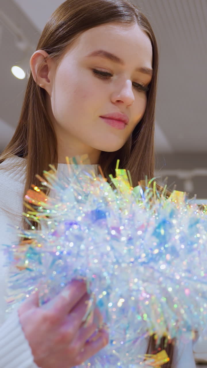 Female shopper holding shimmering tinsel observing it thoughtfully in a decor store, other shoppers, including a child, are seen in the background