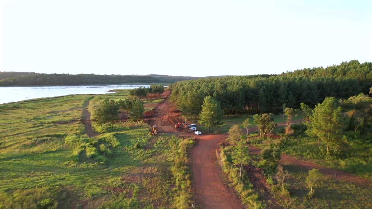 Backward ascending drone clip overwatching rural construction site next to jungle edge and large river, with vast vegetation and distant horizon.