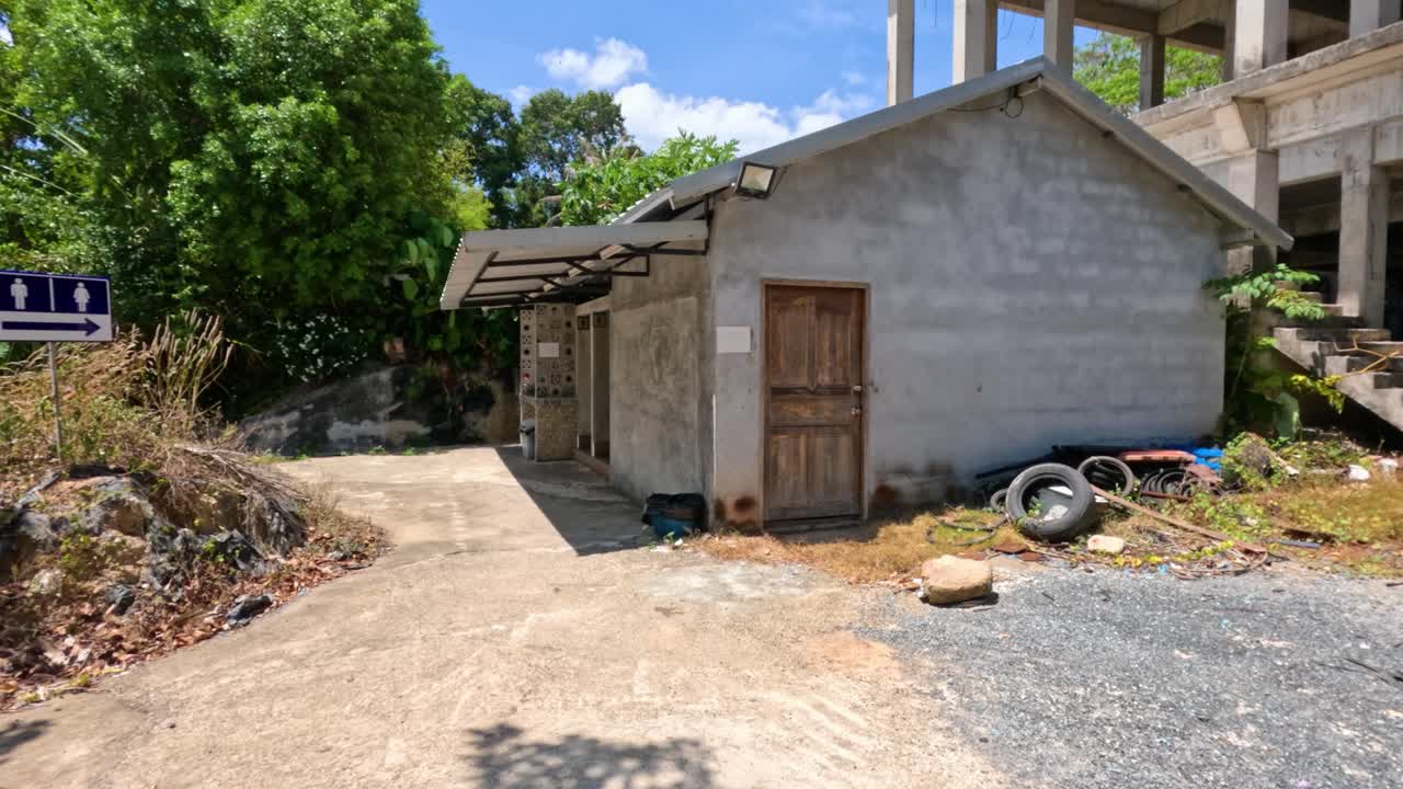 Camera pans past rubbish heap, discarded metal, and tires beside unfinished concrete building, bright daylight