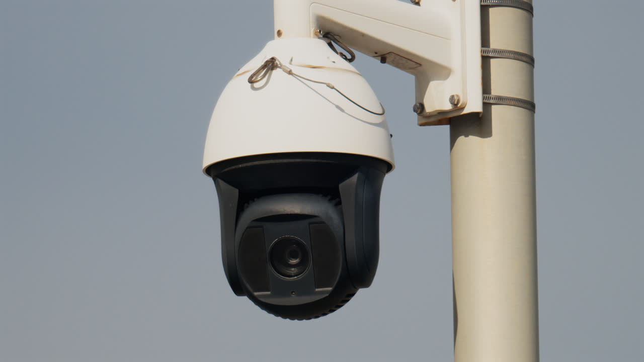 Close up of a round, black and white surveillance camera mounted on a white pole with the cloudy sky on the background
