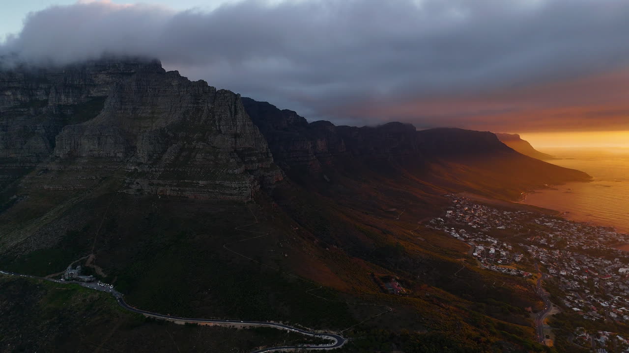 Veduta aerea cinematografica della Table Mountain a Città del Capo, Sudafrica, scogliere rocciose drammatiche con paesaggio urbano costiero al tramonto, nuvole che si muovono sulle cime e luce dorata della sera.