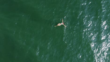 Aerial-view:-Smiling-Latina-in-bikini-swims-alone-in-green-ocean-water