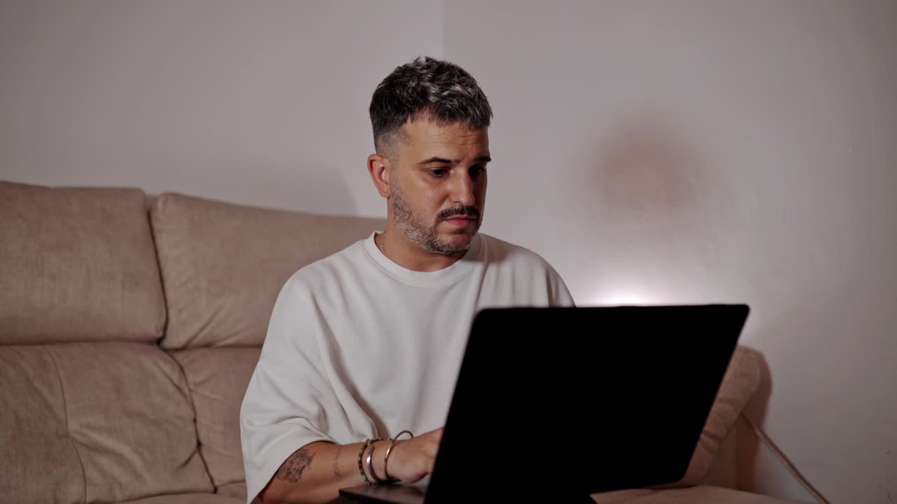 Young man working on laptop with serious expression while sitting on couch at home