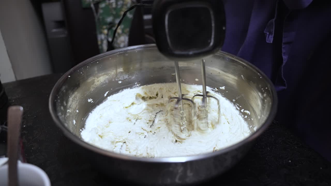 Electric mixer whisking ingredients in a large stainless steel bowl, preparing a dough mixture for baking. slow motion