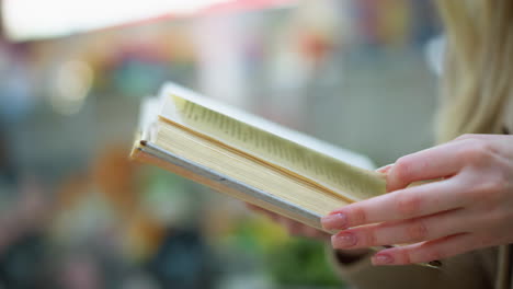 close up hand view of white woman turning to new page in woven gold sweater, modern structure, blur figures and green decorative flower in background