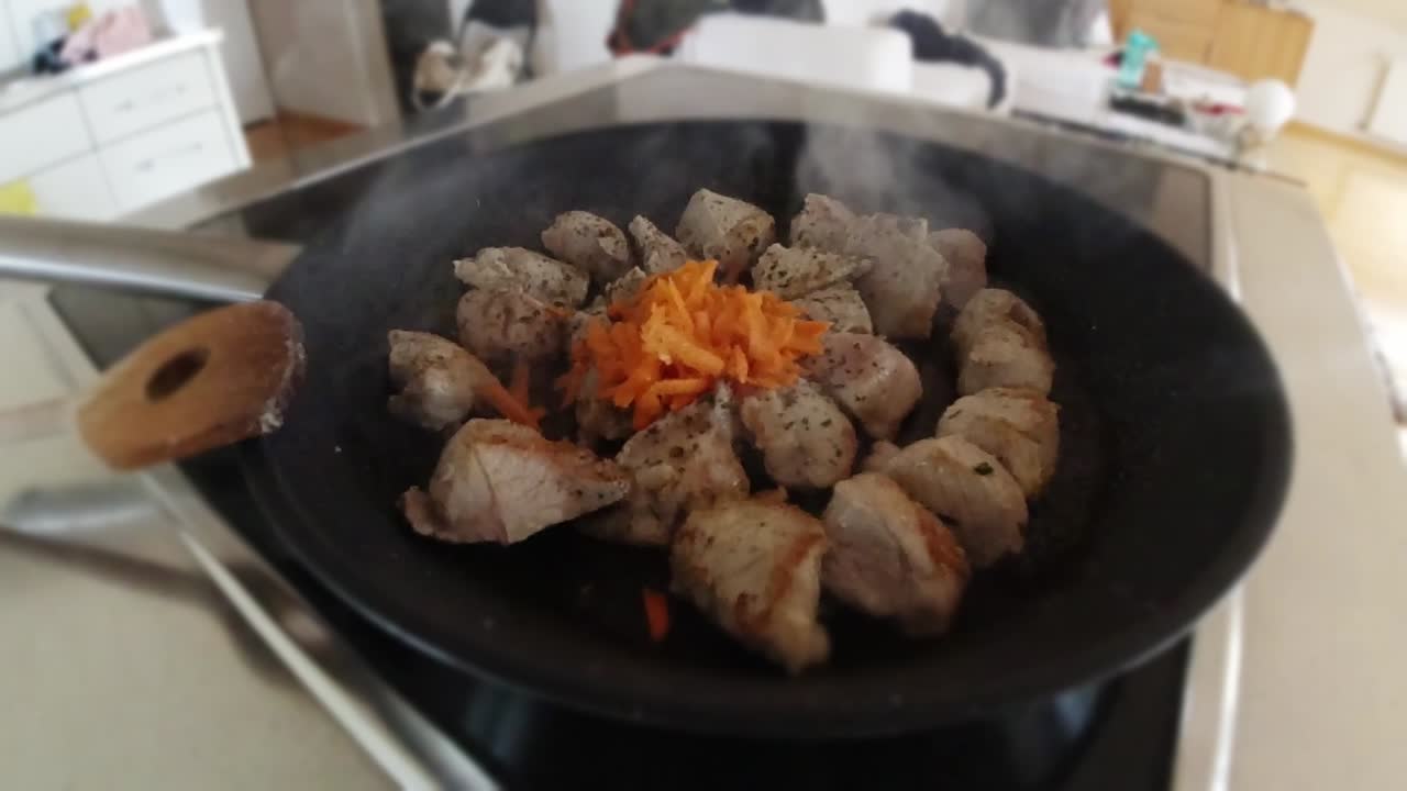 Slow-motion video of chef adding carrots to seared meat in the frying pan in a non-commercial kitchen.