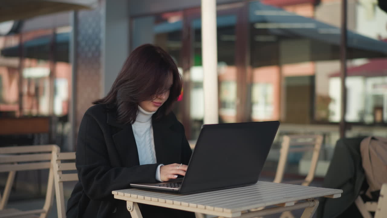 Professional lady focused on laptop screen while seated outdoors in urban environment, modern furniture complements workspace, and background features blurred building