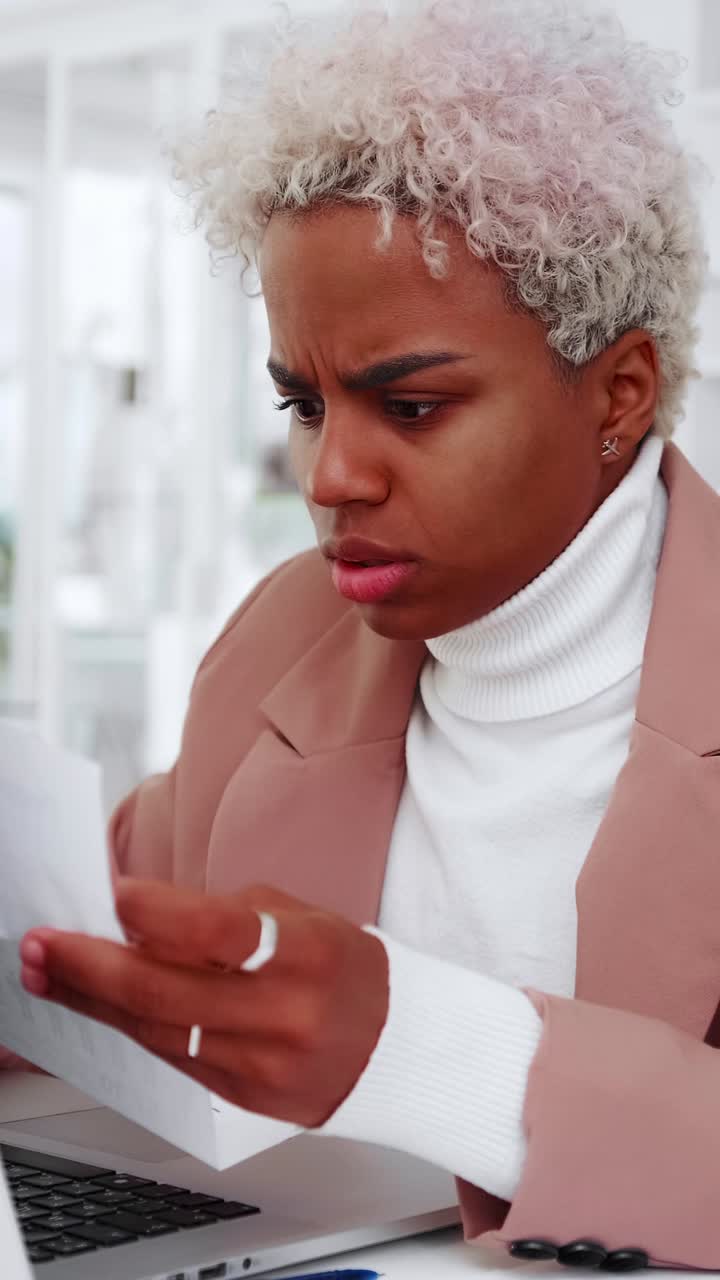 Young woman with focused expression in office reviews printed reports at laptop