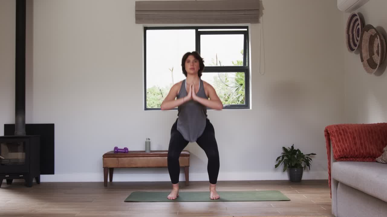 Una donna unisce i palmi delle mani sul tappetino da yoga iniziando una routine di squat sumo alternati ad affondi a casa.