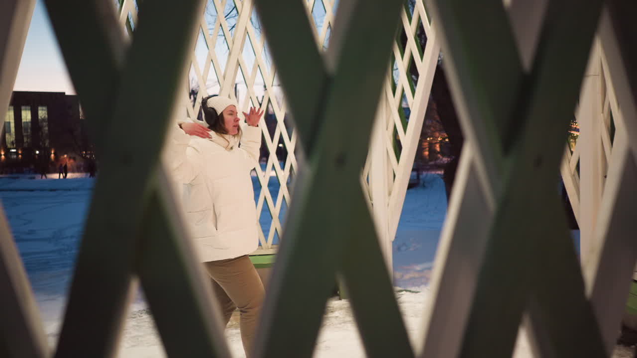Through nets of winter gazebo student wearing white puffer coat and beanie sways body gently to unseen rhythm in snowy park pavilion at dusk under warm lights capturing tranquil seasonal dance mood