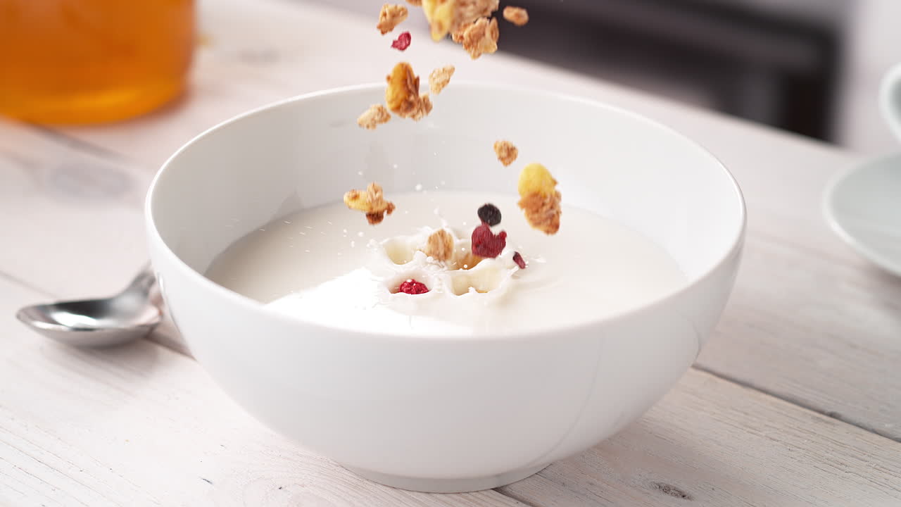 cereales de granola crujientes vertiendo en cámara lenta en un tazón blanco de leche en una mesa de la cocina