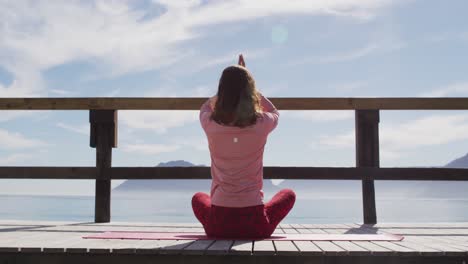 Rear-view-of-of-mixed-race-woman-practicing-yoga-outdoors,-sitting-meditating-by-the-sea