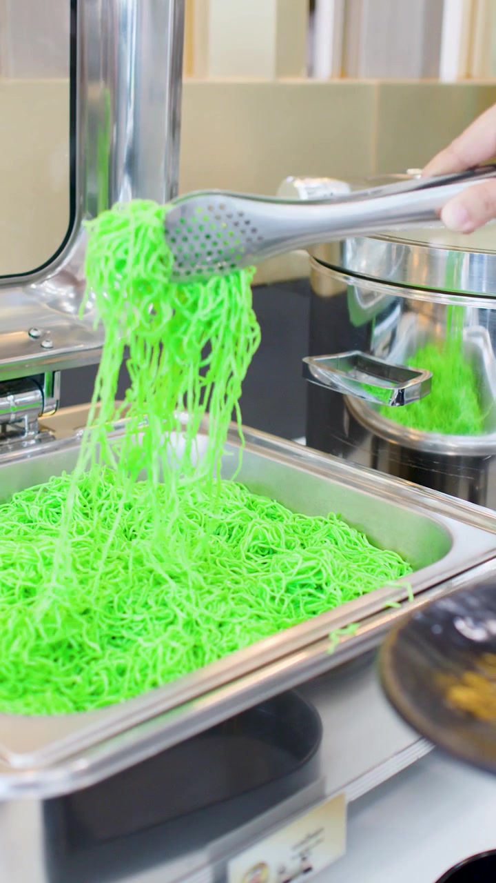 Una persona usa pinze di metallo per servire noodles di giada verde brillante su un piatto nero