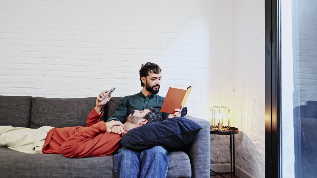 Coppia gay che si rilassa sul divano, uno dei due legge un libro