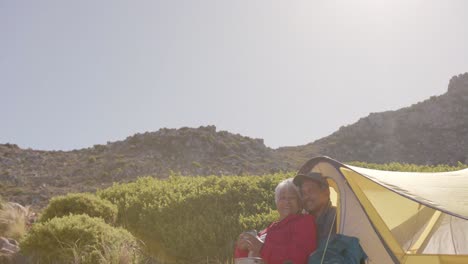 Happy-senior-biracial-couple-sitting-at-tent-in-mountains-and-embracing-on-sunny-day,-in-slow-motion