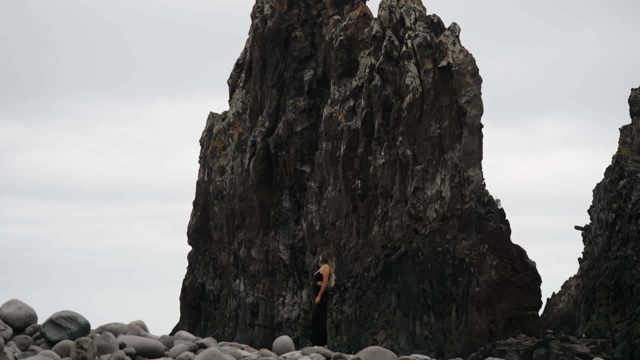A woman stands alone between giant sea stacks and rounded boulders on Madeira’s coastline, dwarfed by the dramatic volcanic cliffs around her.
