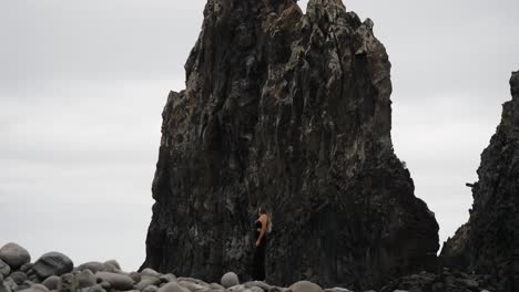 a woman stands alone between giant sea stacks and rounded boulders on madeira’s coastline, dwarfed by the dramatic volcanic cliffs around her.