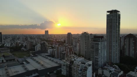 Pan-left-flying-over-Palermo-neighborhood-buildings-and-skyscraper-at-sunset-with-sun-shining-behind-clouds,-Buenos-Aires,-Argentina