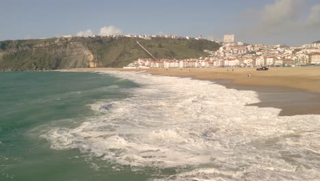 Big-Waves-Lapping-On-The-Seashore-During-Summer-Season-And-Suburb-In-Distance-In-Nazare,-Portugal