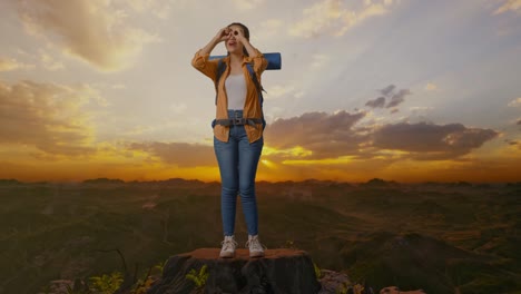 cuerpo lleno de excursionista femenina asiática con mountaineering mochila haciendo binoculares gesto mirando algo y luego diciendo wow mientras está de pie en la cima de la montaña durante la hora de la puesta del sol