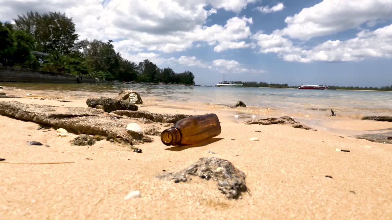 Brown glass bottle and coral fragments on sandy beach, bright daylight, slight camera movement, Phuket