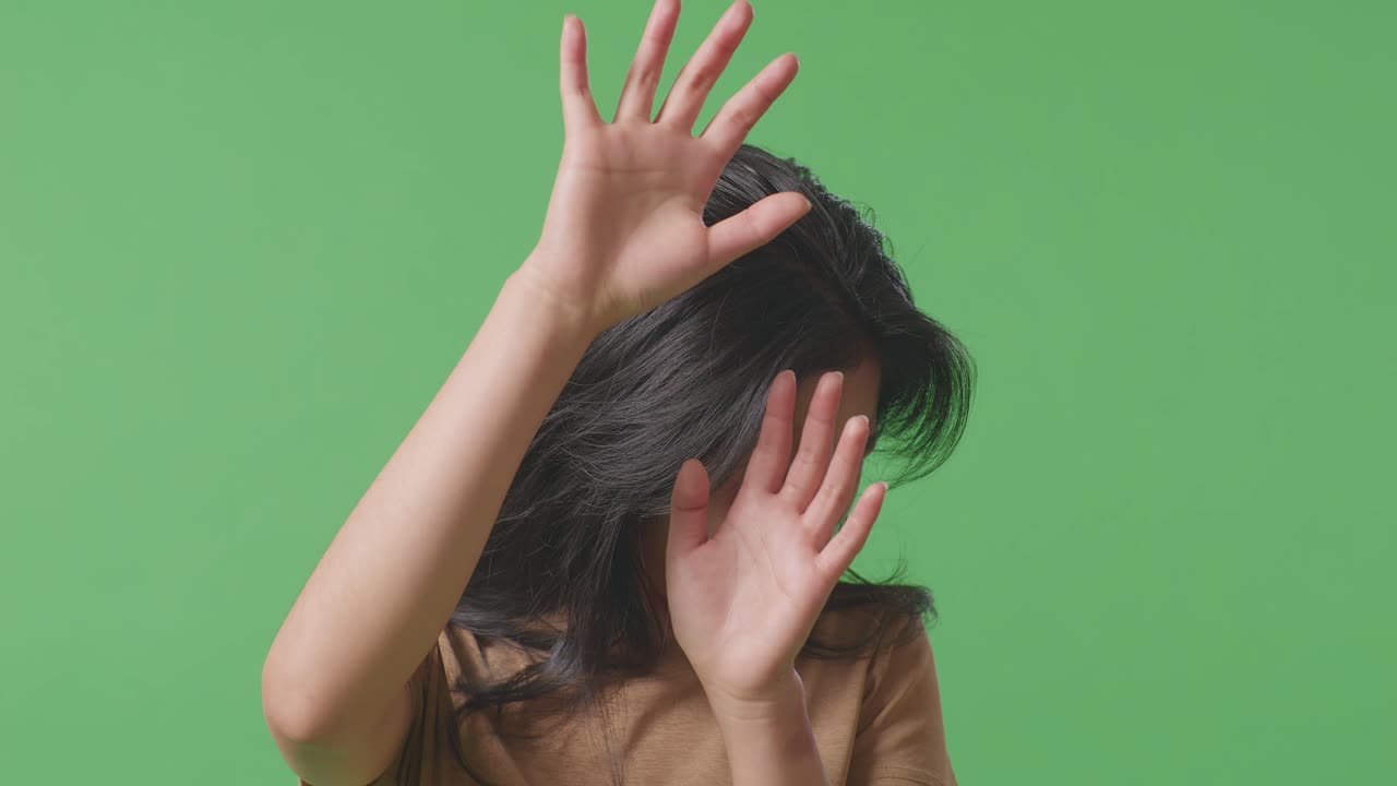 얼굴에 이 있는 폭력의 희생자인 젊은 아시아 여성이 녹색 스크린 스튜디오에서 손으로 방어하는 몸짓을 하고 두려워합니다.